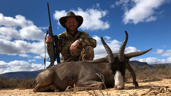 Hunter poses with a downed animal against a blue cloud-filled sky