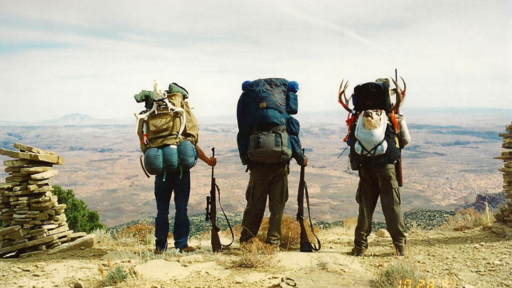 Three hunters with packs on gaze off mountainside with backs to camera