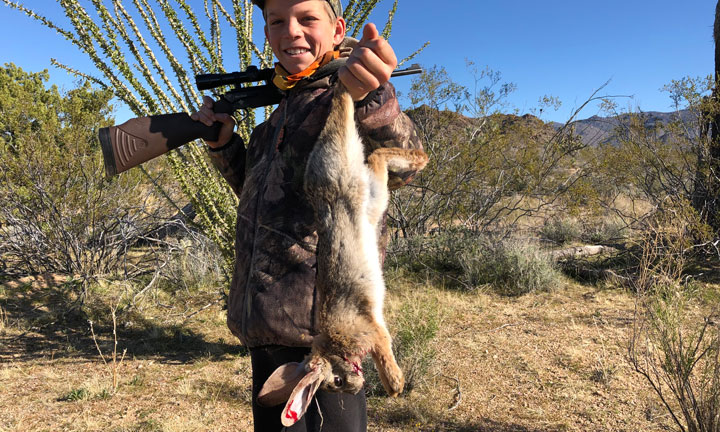 Young hunter in the scrublands holding a dead rabbit by its hind legs, with a rifle over his shoulder.