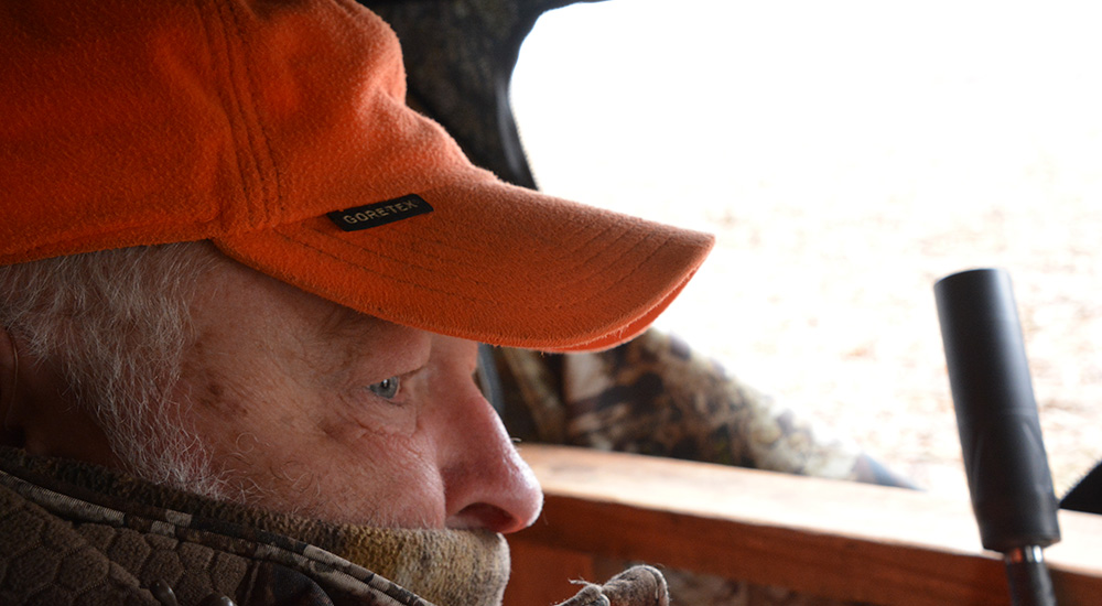 Male hunter with blaze orange hat sitting in hunting blind.