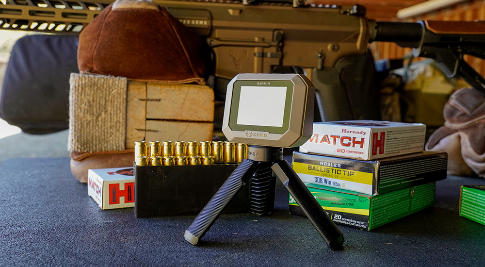 Garmin handheld chronograph at shooting range next to rifle and boxes of ammunition.