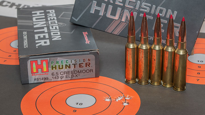 A five shot grouping of Hornady measures to less than an inch. Target is orange an dblack, five cartridges tand up to the right of the photo, and two black boxes of Hornady Precision Hunter are propped in the back ground.