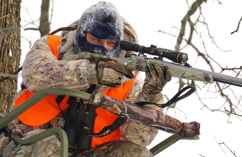 Hunter shouldering rifle while sitting in deer stand.