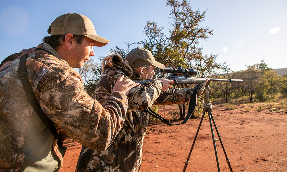 Female hunter shooting bolt action rifle off sticks with African professional hunter behind.