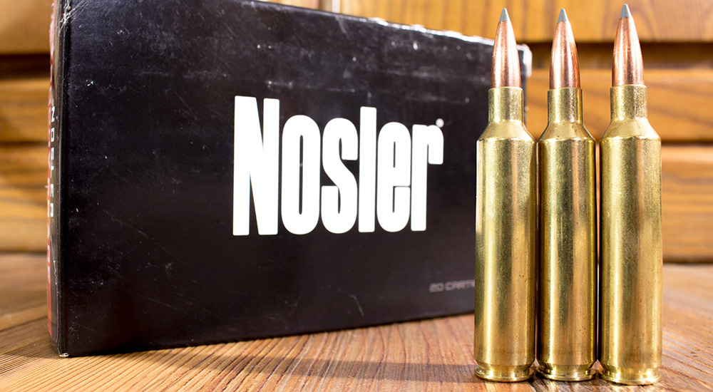 Nosler ammunition box with three 27 Nosler ammunition rounds standing on wood table.