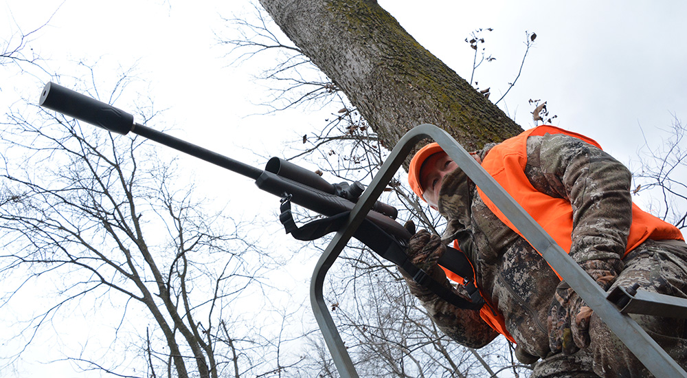 Male hunter in tree stand with rifle resting on safety bar.