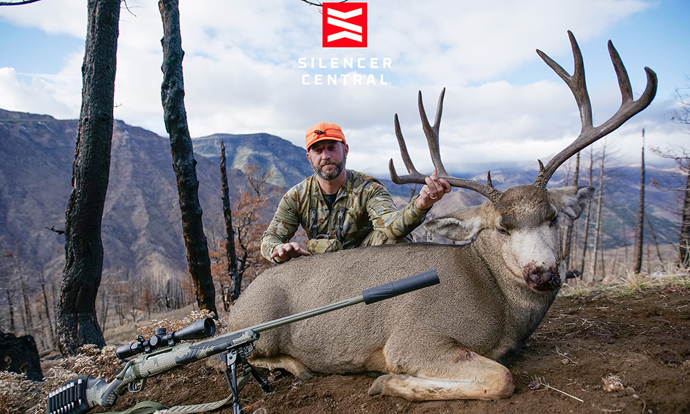 Male hunter posing with mule deer buck with bolt action rifle fitted with suppressor resting on ground.