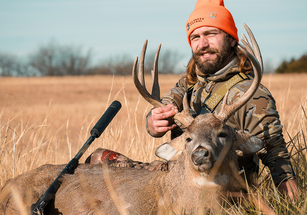 Young adult male hunter posing with whitetail buck in Kansas field.