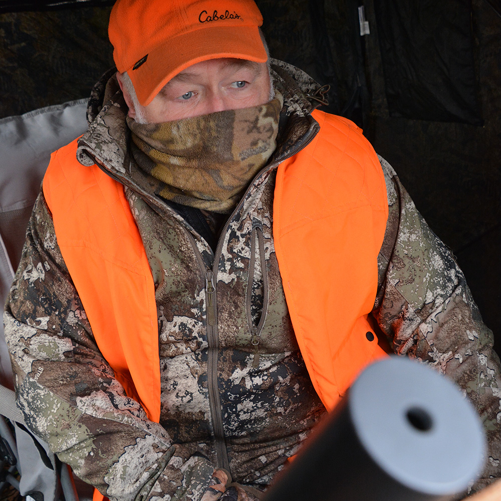 Male hunter sitting in hunting blind with camouflage and blaze orange vest and hat.