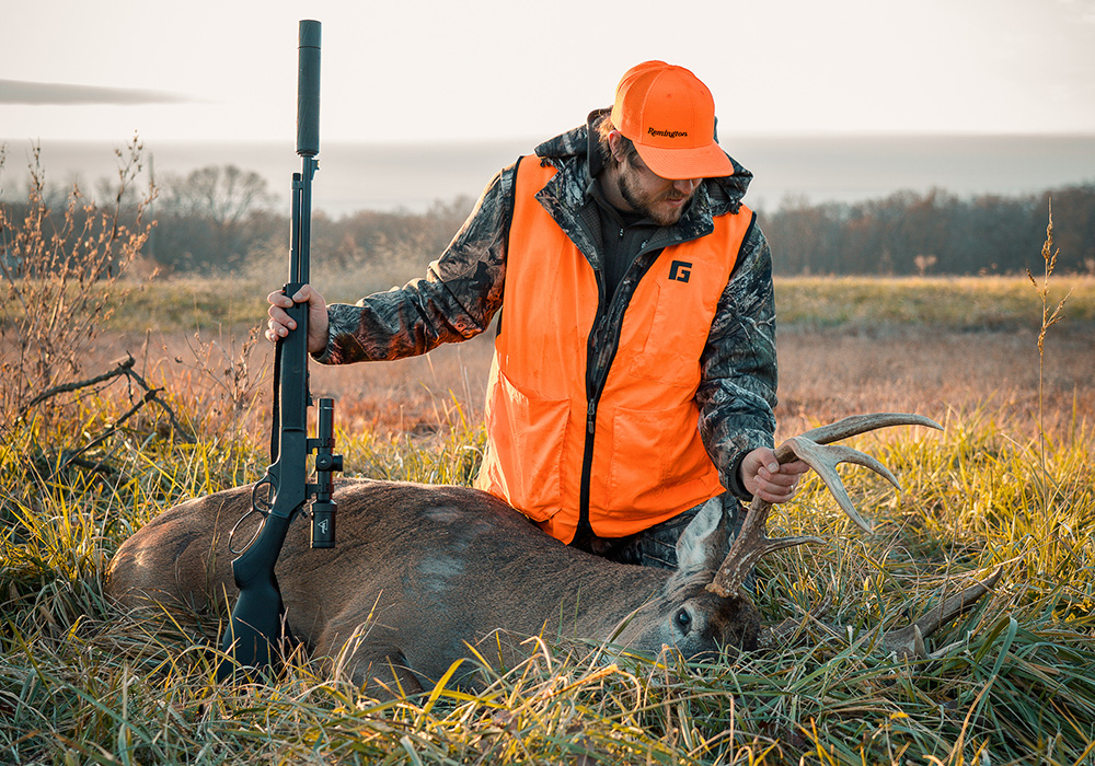 Male hunter kneeling over whitetail buck in field while holding rifle.