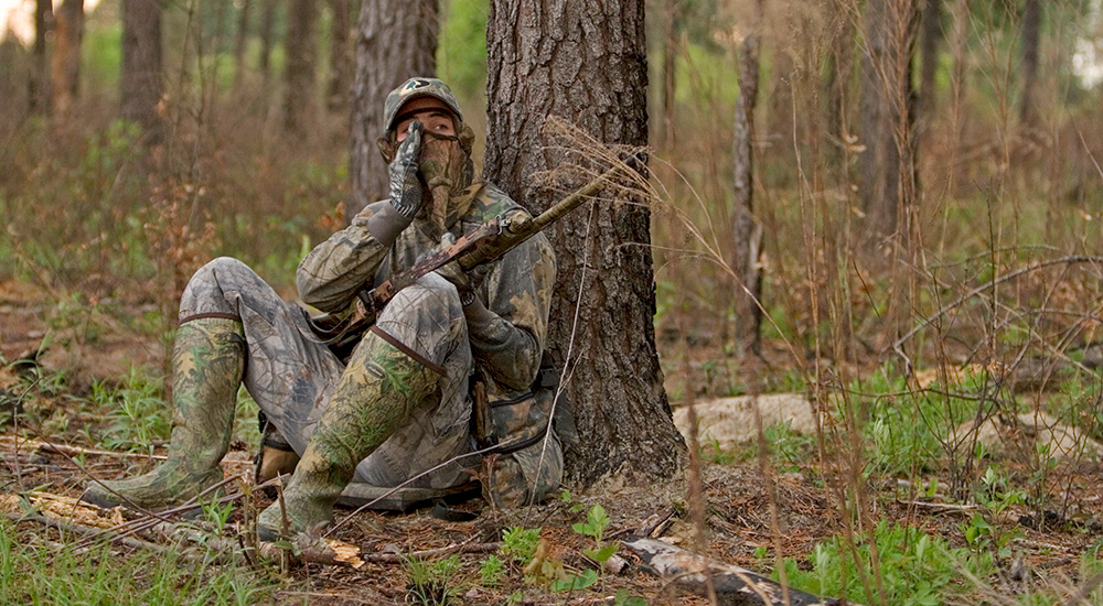 Turkey hunter sitting at base of tree in woods.